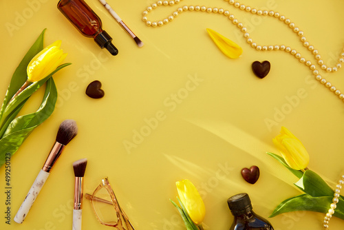  Trendy yellow feminine flat lay: makeup brushes, glasses, serum dropper, pearls, chain. Golden accessories, chocolate hearts and yellow tulips. Beauty card concept. Copy space photo
