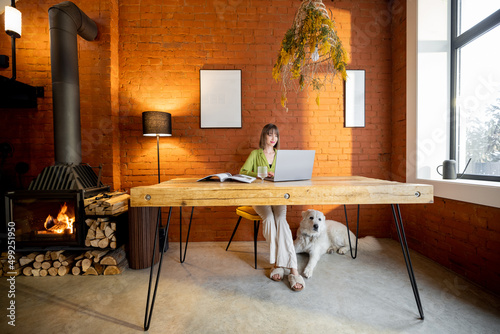 Woman works on laptop while sitting with her dog by the wooden table in cozy living room with a burning fireplace. Interior in loft style photo