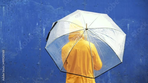 Unrecognizable woman in yellow raincoat under rain spinning around an umbrella. Horizontal rear view of female outdoors twirling around transparent umbrella on blue background. Weather concept