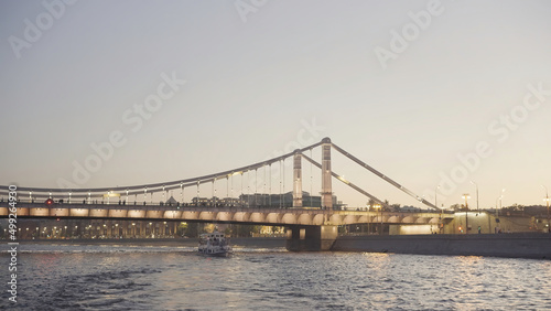 Beautiful city landscape with river glowing bridge and floating boat. Action. Boat floats on river on background of glowing bridge at sunset