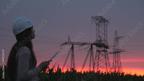 Power engineer woman works in protective helmet checks power line using data of computer tablet. Energy crisis. High voltage power lines at sunset. Distribution and supply of electricity. Clean energy