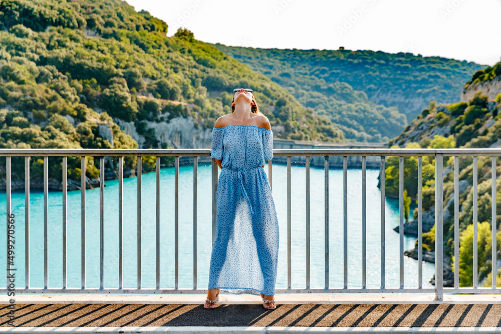 The beautiful girl in a blue dress and sunglasses poses on the bridge, the long chestnut hair, happy and smiles, azure water of the lake and slopes of mountains on a background