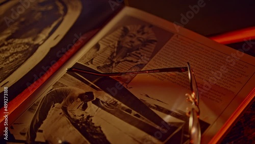 Camera racking focus across rare ancient Egyptian archaeology book. Black and white photo of Tutankamen's death mask and Howard Carter excavation featured. Cinematic lighting.