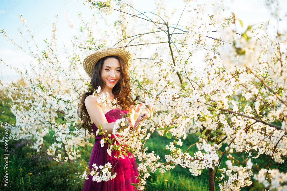 Portrait of a dreamy girl in the spring park among the flowering white trees at sunset. Woman in straw hat and purple dress, cherry blossoms.