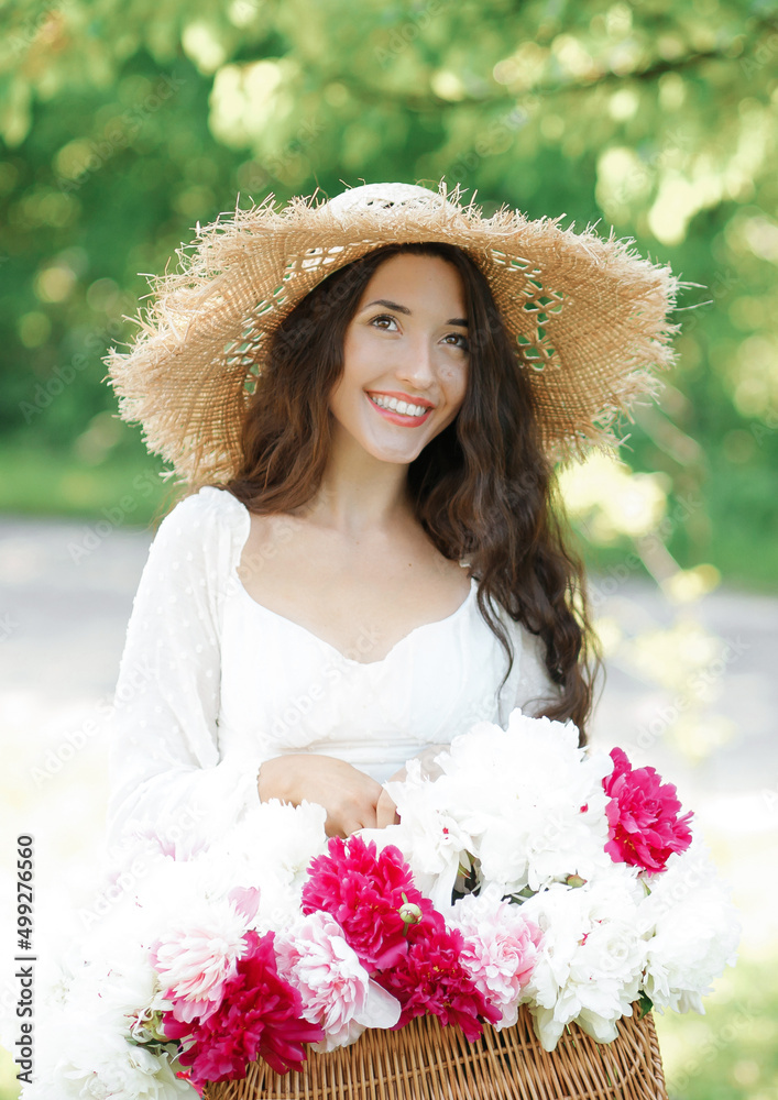 Happy smiling girl in a straw hat holding a wicker basket with white and pink peony flowers posing on the streets of a European city. Lifestyle