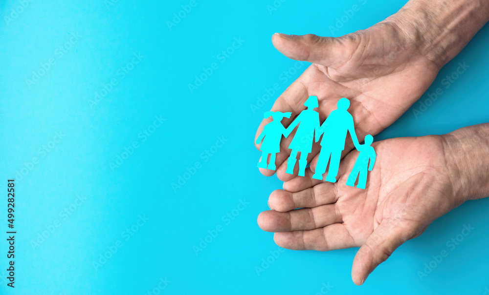 The hands of an elderly person hold a paper-cut silhouette of a family on a blue fog. Copyspace on the left. Family day, family traditions concept. Family tree, succession of generations. The hands of an elderly person hold a paper-cut silhouette of a family on a blue fog. Copyspace on the left. Family day, family traditions concept. Family tree, succession of generations.