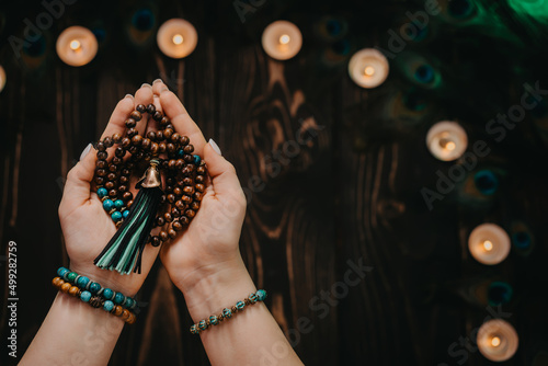 Woman holds in hand wooden mala beads strands used for keeping count during mantra meditations. Weaving and creation. Wooden background with candles and feathers. Spirituality, religion, God concept.