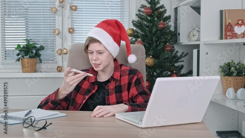 A cheerful teenage boy in a Santa hat, chatting on a mobile phone, sitting at a computer table indoors at home, smiling, enjoying a conversation.