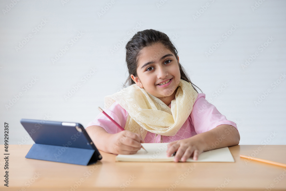 portrait cute Indian girl writing on notebook and online learning class from tablet, education concept portrait cute Indian girl writing on notebook and online learning class from tablet, education concept