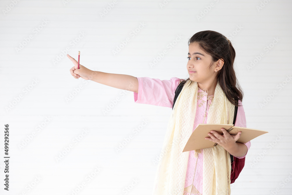 cute Indian girl holding notebook and pencil, pointing to something on white wall background cute Indian girl holding notebook and pencil, pointing to something on white wall background