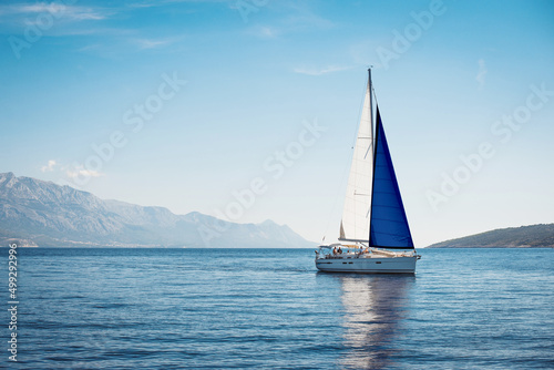A white yacht with sails the color of the Finnish flag in the sea against a background of blue sky and mountains © Maxim Sokolov