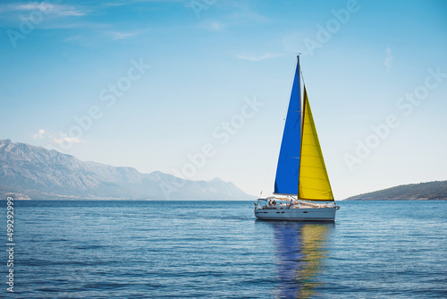 A white yacht with sails the color of the Ukrainian flag in the sea against a background of blue sky and mountains © Maxim Sokolov