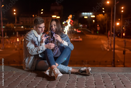 Couple of students in love are sitting on evening city background with garland in their hands. Night city lights