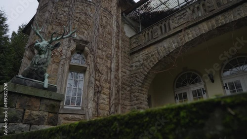 Tilt shot at the entrance of an old castle, revealing deer statue and an ivy-clad tower photo