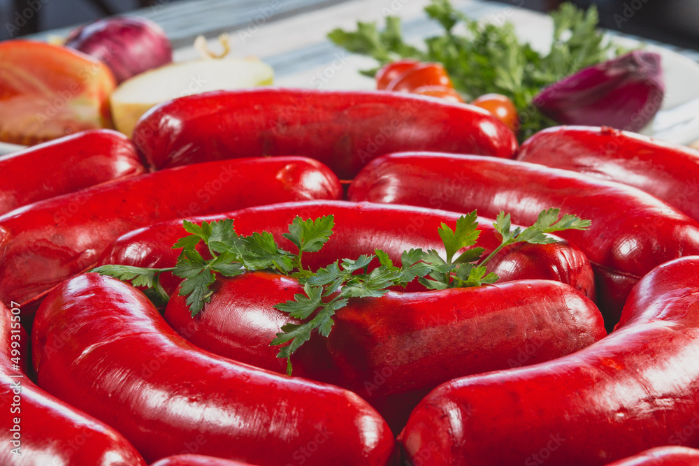 Close Linguiça calabresa vermelha grande crua em gomos sobre tábua de madeira em toalha azul e cinza acompanhado de tomate cereja cebola roxa alface salsa in natura cortados