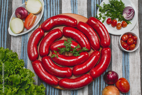 Fototapeta Naklejka Na Ścianę i Meble -  Linguiça calabresa vermelha grande crua em gomos sobre tábua de madeira em toalha azul e cinza acompanhado de tomate cereja cebola roxa alface salsa in natura cortados