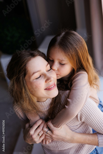Lovely family mother and daughter embracing and kissing each other indoors. Happy sweet woman with little 6-year-old girl in casual clothers: turtlenecks and jeans sitting on sofa, hugging and smiling