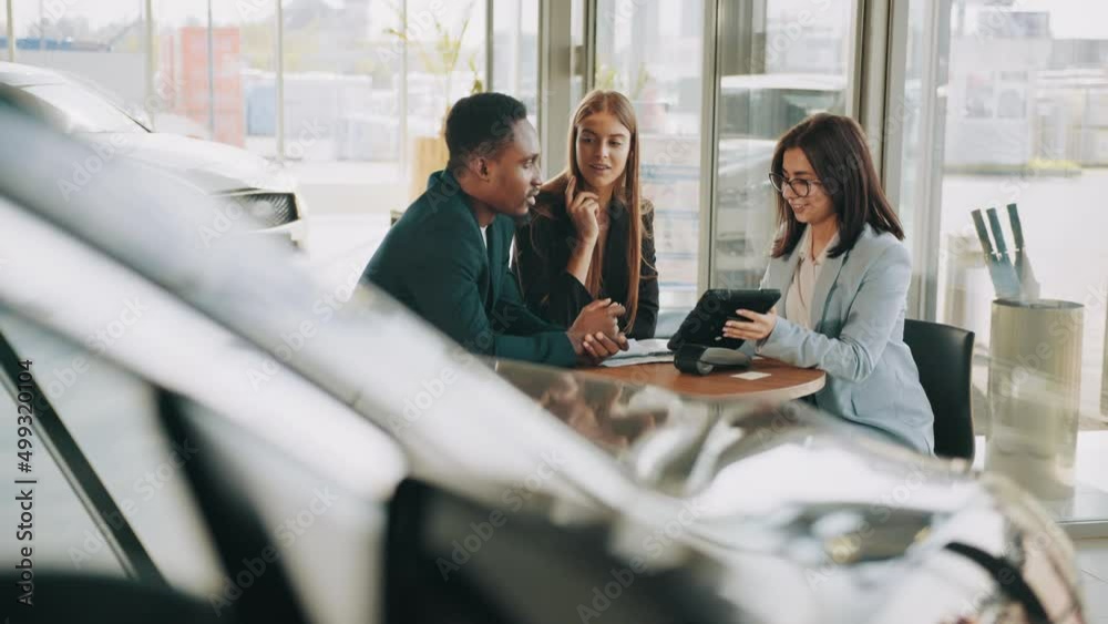 Cheerful multicultural couple sitting with saleswoman at modern salon. Successful agreement of car purchsing. Auto business concept. Manager using digital tablet while working with buyers at showroom