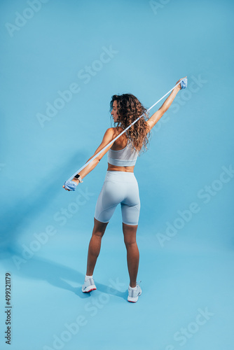 Rear view of fit young woman stretching arms with rubber band on blue background