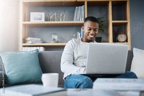 Im having the best time all by myself. Shot of a handsome young man using his laptop while sitting on a sofa at home.