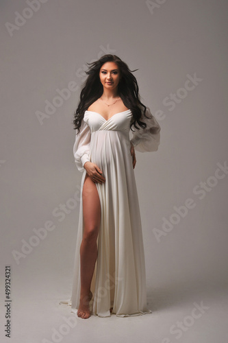 Portrait of attractive brunette pregnant woman in white dress holding her pregnant belly, isolated on cyclorama. The concept of happy motherhood and parenthood
