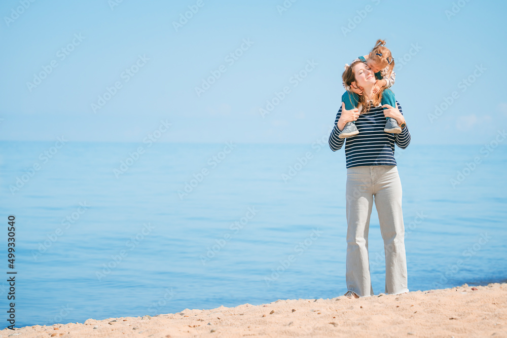 Daughter sits on mother's shoulders, hugs and kisses mom on the beach on sunny day. Happy family on the beach
