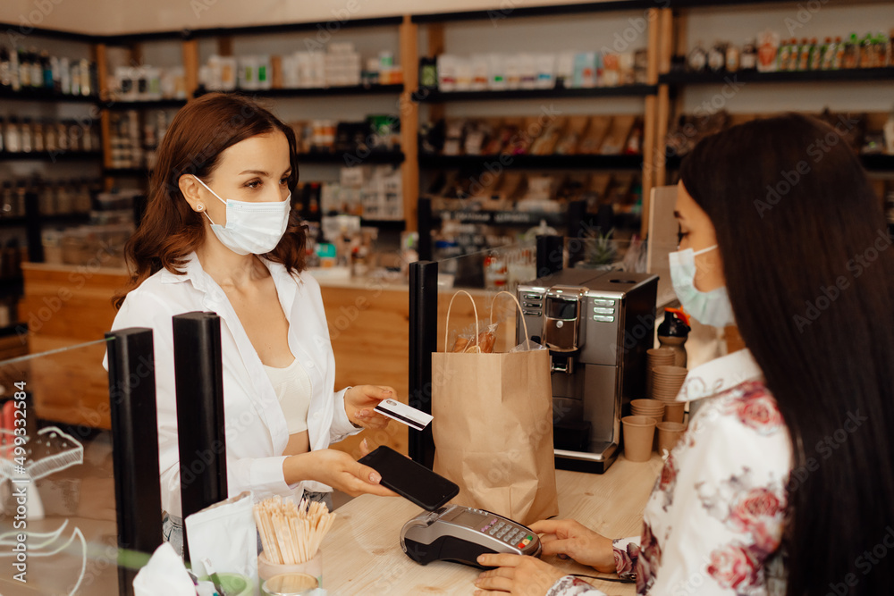 Girl buyer in a mask makes a purchase in the store. Saleswoman or small business owner wearing medical mask at the counter in cafe or small shop. Girl buyer in a mask makes a purchase in the store. Saleswoman or small business owner wearing medical mask at the counter in cafe or small shop.