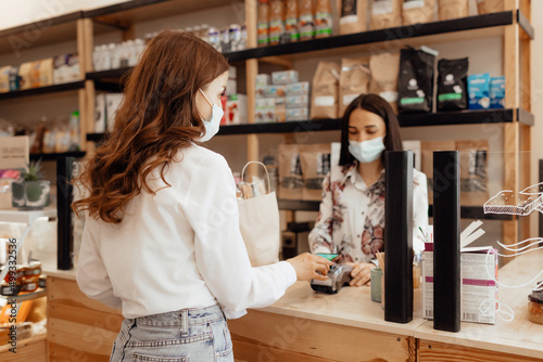 Girl buyer in a mask makes a purchase in the store. Saleswoman or small business owner wearing medical mask at the counter in cafe or small shop.