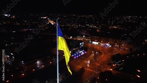 Illuminated flag of Ukraine waving on dark night cityscape. City counterclockwise aerial view on traffic on Pavlivska Square near Skver Strilka in Kharkiv downtown, Ukraine photo