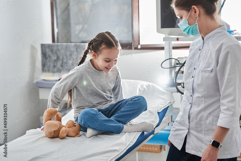 Doctor wearing protective mask standing near the hospital bed and talking with her little patient Doctor wearing protective mask standing near the hospital bed and talking with her little patient
