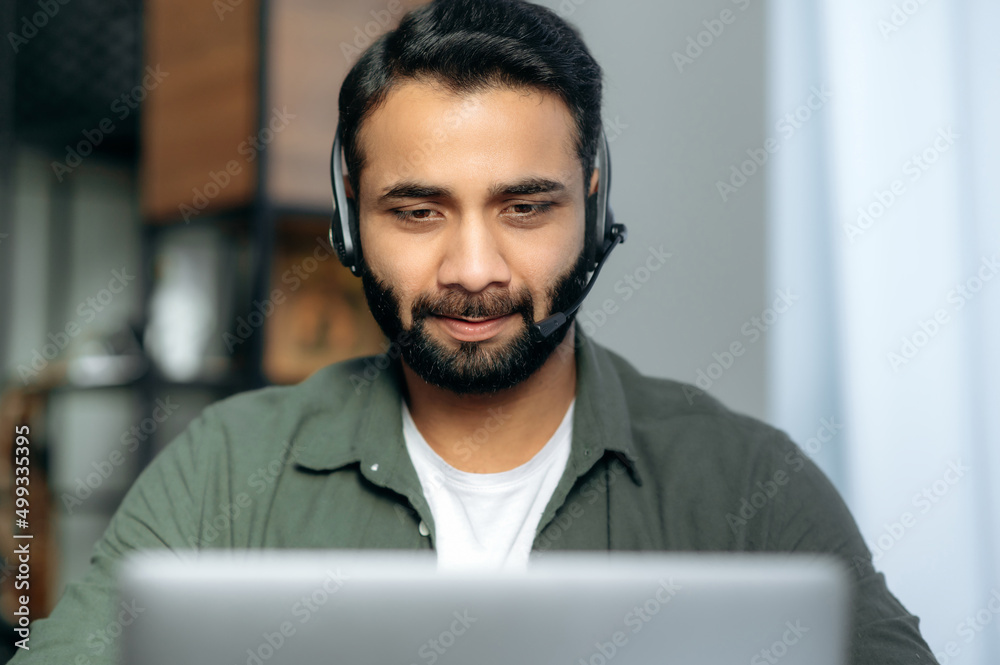 Close-up of focused serious Indian or Arabian man wearing headset, expert, consultant or mentor, looks at laptop screen, conducts online consultation. Support service concept, hotline Close-up of focused serious Indian or Arabian man wearing headset, expert, consultant or mentor, looks at laptop screen, conducts online consultation. Support service concept, hotline