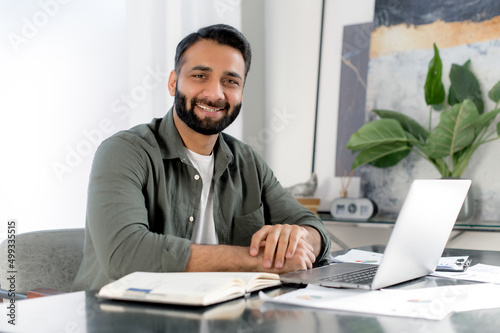 Positive confident indian or arabian man using laptop in modern office. Portrait successful businessman, freelancer, manager, sits at workplace in front of laptop, looking at camera, smiling friendly