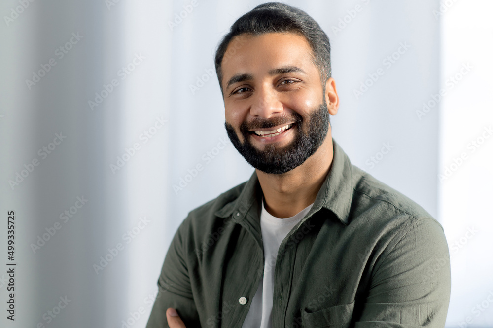 Obraz premium Portrait of indian or arabian positive successful businessman, manager, freelancer, office worker, wearing casual stylish shirt, stand in an modern office, smiling friendly at the camera