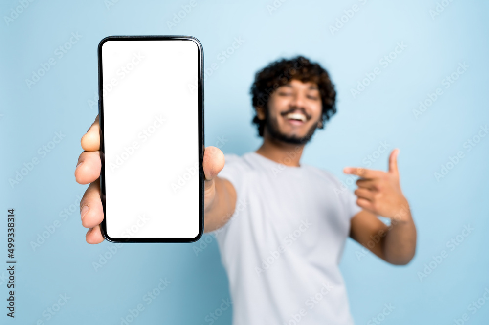 Mock-up, copy space. Defocused indian or arabian guy, in casual wear, holds smartphone with blank white screen for presentation and points finger at it, stands on isolated blue background, smiling