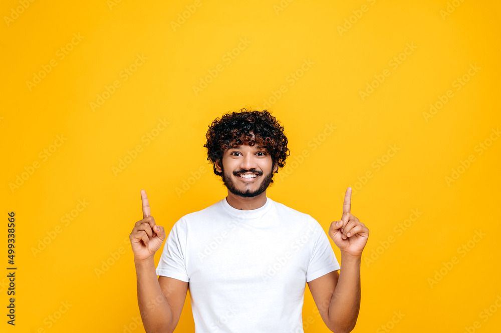 Positive excited indian or arabian guy in basic white t-shirt, amazed looks at the camera and points fingers up, at empty mock-up space, stands on isolated orange color background. Mock-up concept