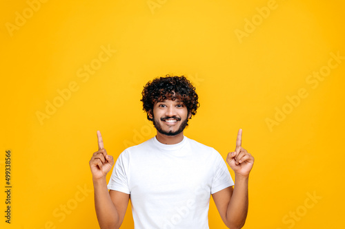 Fototapeta Positive excited indian or arabian guy in basic white t-shirt, amazed looks at the camera and points fingers up, at empty mock-up space, stands on isolated orange color background