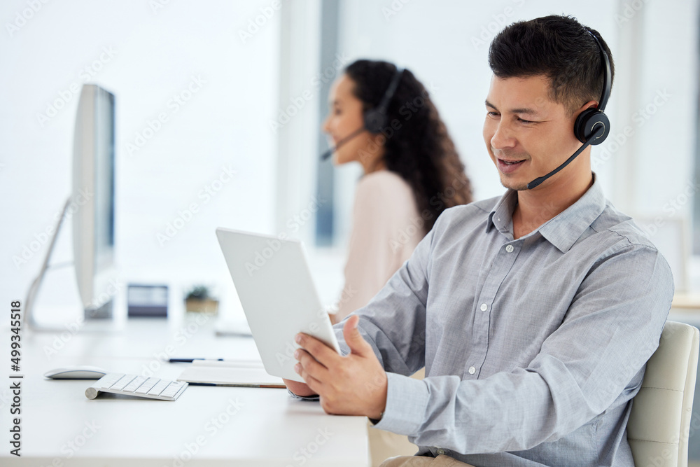 Hes such a smart salesman. Shot of a young call centre agent working on a digital tablet in an office with his colleague in the background. Hes such a smart salesman. Shot of a young call centre agent working on a digital tablet in an office with his colleague in the background.