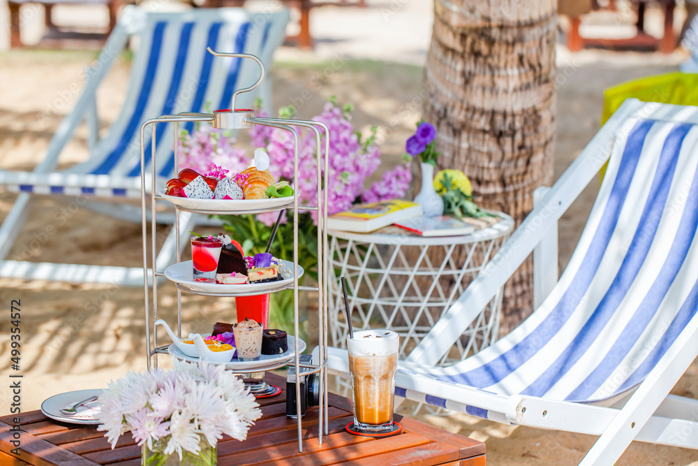 Afternoon tea on tropical sandy beach with palm trees and sun lounges on background. Cake stand with fresh pastries, sweeties, iced coffee and shake. Travel in Thailand, luxury hotel and restaurant