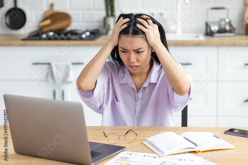 Billede på lærred Young Asian woman freelancer, looking worried at laptop screen, holding her head