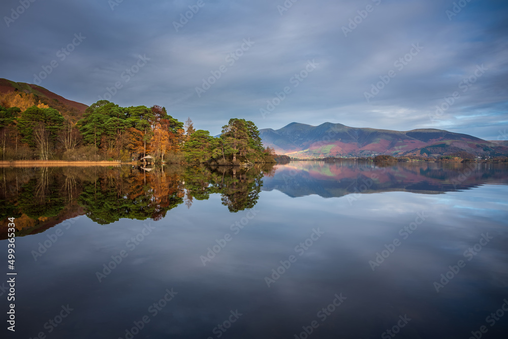 Absolutely stunning vibrant Autumn sunrise landscape image looking from Manesty Park in Lake Distict towards sunlit Skiddaw Range with mit rolling across Derwentwater surface