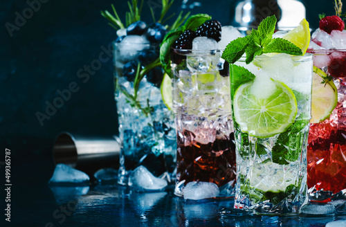 Cold summer cocktails drinks. Classic alcoholic long drink and mojito or mocktail in highballs with blueberries, blackberries, raspberries, lime, herbs and ice on blue background