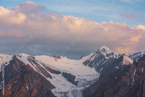 Dramatic aerial view to high snow mountain peak in early morning at dawn. Awesome scenery with sunlit snow mountains in cloudy sky at sunrise. Scenic landscape with large glacier in sunrise colors.
