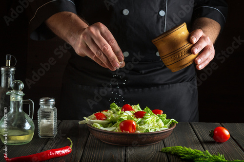 Tableau sur toile The cook sprinkles salt to salad of fresh vegetables on wooden table