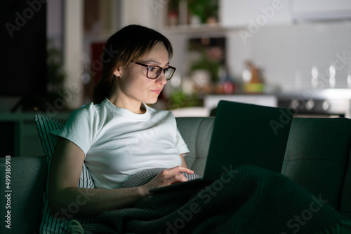 Serious young woman in glasses using laptop sitting on couch at home in the night. Thoughtful girl freelancer watching education video, lying on sofa and working on computer in dark room. 