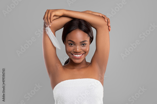 Smiling pretty millennial black female with perfect skin showing armpits and hold feather, isolated on gray background