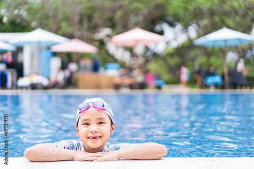 Asian child smile toothless or kid girl wearing swimsuit and cap on edge swimming pool and happy fun in waterpark for learning swim to exercise on summer school or vacation travel with copy space © kornnphoto