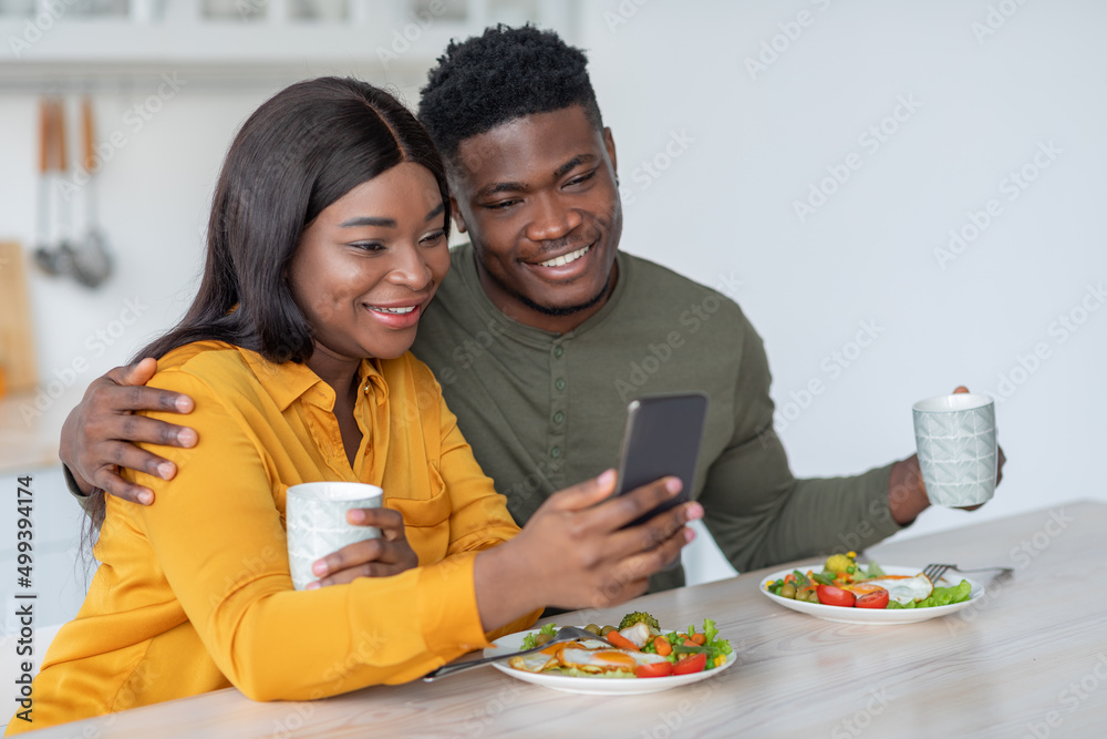 Young Black Man And Woman Using Smartphone While Enjoying Breakfast In Kitchen