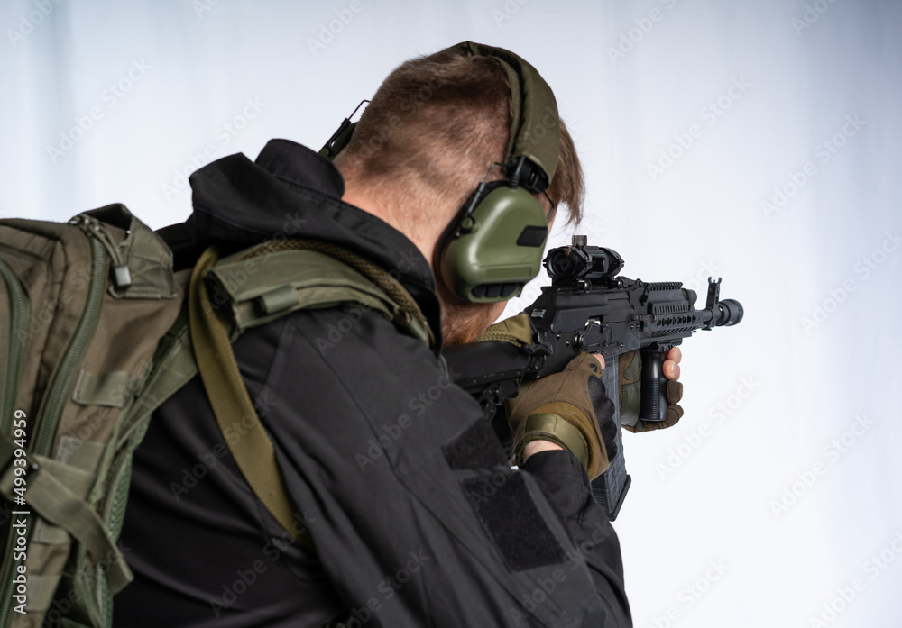 an airsoft soldier in full gear with his back to us is aiming at an optical sight. a man in an outfit, in headphones, a bulletproof vest, with a backpack and a belt. White background.