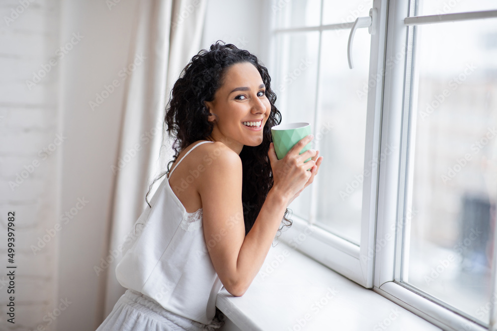 Glad young caucasian brunette lady with long hair in pajamas drink favorite hot tea near window in bedroom Glad young caucasian brunette lady with long hair in pajamas drink favorite hot tea near window in bedroom