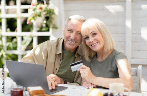 Online shopping concept. Modern senior couple using laptop and credit card, sitting outdoors in countryside